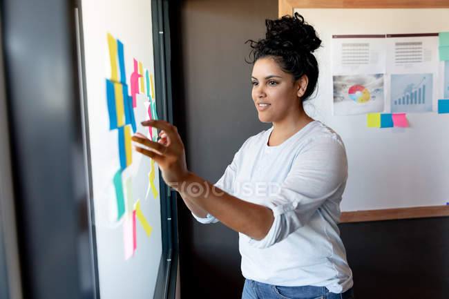 Vue latérale d'une jeune femme métisse travaillant dans un bureau créatif debout près d'un tableau blanc mettant des notes de service sur . — Photo de stock