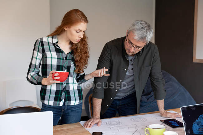Vista frontal de uma jovem mulher caucasiana e um homem caucasiano trabalhando em um escritório criativo, em pé ao lado de uma mesa, olhando para os planos arquitetônicos . — Fotografia de Stock
