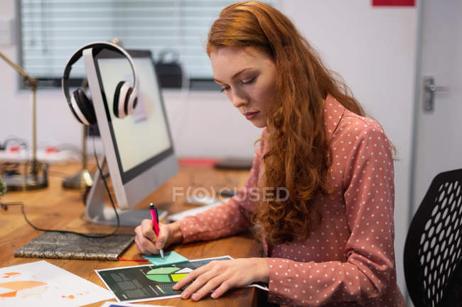 Vista lateral de uma jovem mulher caucasiana trabalhando em um escritório criativo, escrevendo e sentado em uma mesa com um computador ligado — Fotografia de Stock