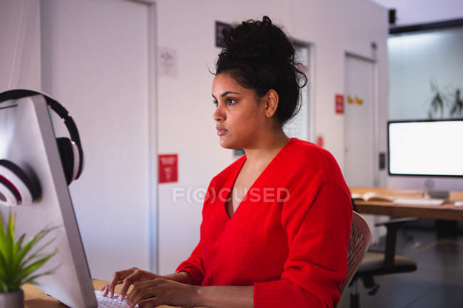 Vista lateral de uma jovem mulher de raça mista trabalhando em um escritório criativo, sentado em uma mesa usando um computador, digitando — Fotografia de Stock
