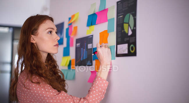 Vue latérale d'une jeune femme caucasienne travaillant dans un bureau créatif debout près d'un tableau blanc écrivant sur des notes de service — Photo de stock