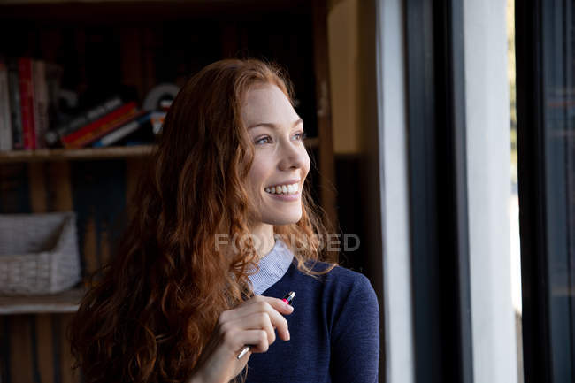 Vue latérale d'une jeune femme caucasienne heureuse dans un bureau créatif, souriante et regardant par la fenêtre . — Photo de stock