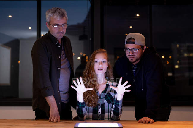 Вид спереди на молодую кавказку и двух белых мужчин за столом в креативном конференц-зале, на встрече, за планшетным компьютером, глядя на экран, женщина жестикулирует руками . — стоковое фото