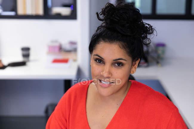 Porträt einer jungen Frau mit gemischter Rasse, die an einem Schreibtisch in einem kreativen Büro sitzt und in die Kamera lächelt. — Stockfoto