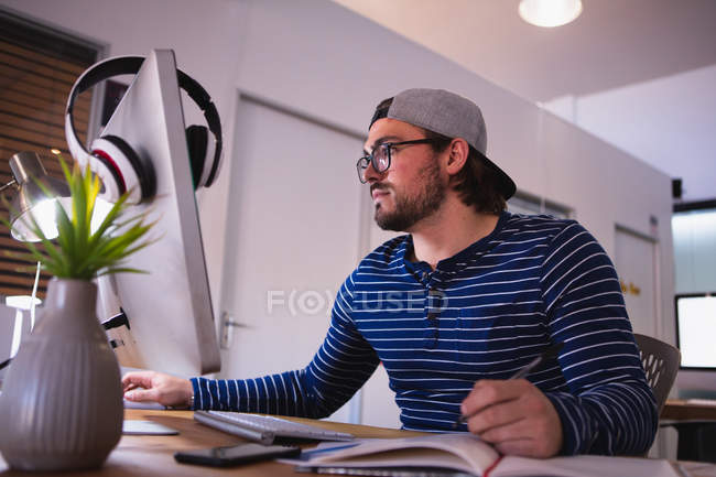 Vista lateral de um jovem caucasiano trabalhando em um escritório criativo, sentado em uma mesa usando um computador e escrevendo, usando óculos e boné — Fotografia de Stock