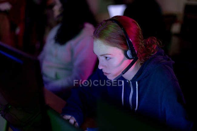 Side view of a young gamer Caucasian woman working in a creative office with headset on looking at computer screen with her female colleague in the background — Stock Photo