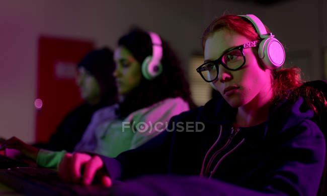 Вид збоку на молоду кавказьку жінку-гравця, що працює в творчому офісі, носить окуляри, слухає музику з навушниками, а її колеги працюють на задньому плані — стокове фото