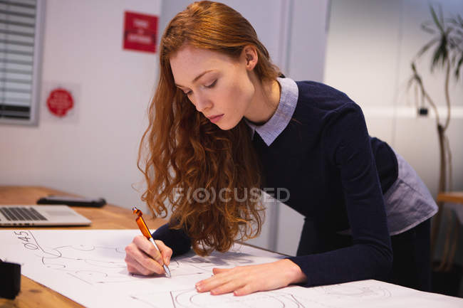 Side view of a young Caucasian woman working in a creative office, leaning on her desk and making notes on paper — Stock Photo