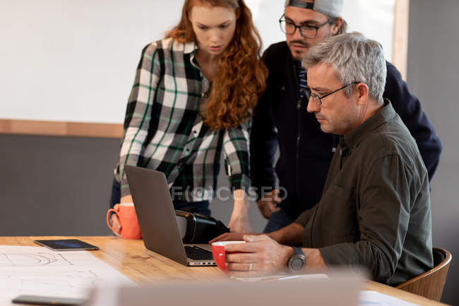Vista frontale di una giovane donna caucasica e due uomini caucasici che lavorano in un ufficio creativo da una scrivania, usando un computer portatile e guardando lo schermo . — Foto stock