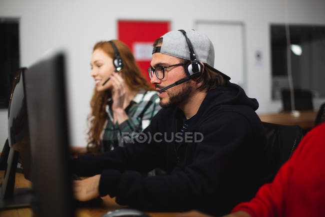 Вид сбоку на молодого кавказского геймера, работающего в креативном офисе, в очках для чтения и наушниках, смотрящего на экран компьютера со своей коллегой-женщиной на заднем плане — стоковое фото