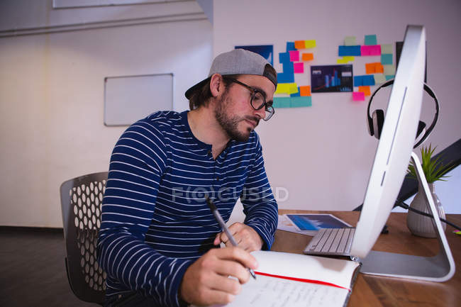 Side view of a young Caucasian man working in a creative office, sitting at a desk writing and looking at computer screen, wearing glasses and cap — Stock Photo