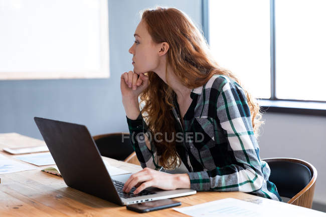 Vista lateral de uma jovem mulher caucasiana trabalhando em um escritório criativo, usando um computador portátil . — Fotografia de Stock