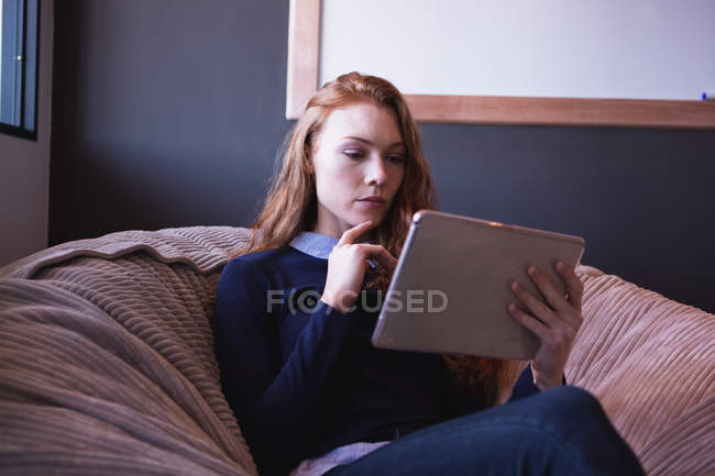 Front view of a young Caucasian woman working in a creative office, holding a tablet computer and sitting on a bean bag — Stock Photo