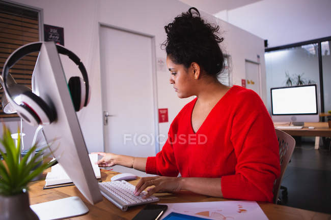 Вид збоку молода змішана жінка, що працює в творчому офісі, сидить за столом за допомогою комп'ютера — стокове фото