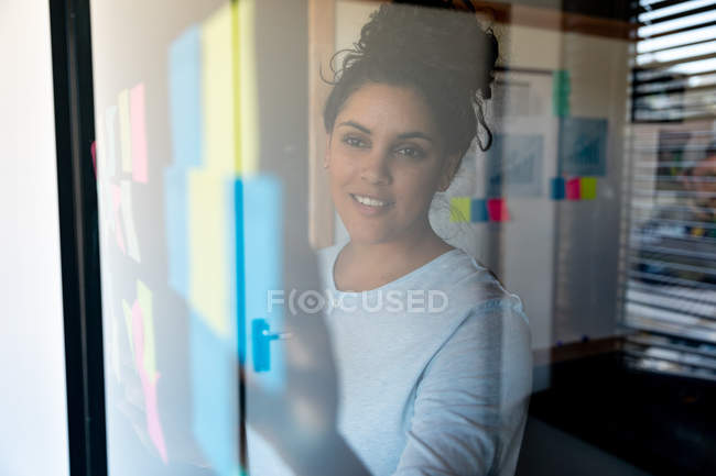 Vista frontale di una giovane donna di razza mista che lavora in un ufficio creativo in piedi vicino a una finestra, mettendo appunti e sorridendo . — Foto stock