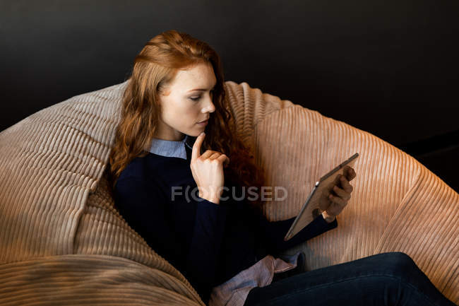 Vista lateral de uma jovem mulher caucasiana trabalhando em um escritório criativo, segurando um computador tablet e sentado em um saco de feijão . — Fotografia de Stock