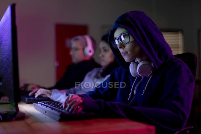 Vista lateral de una joven jugadora caucásica que trabaja en una oficina creativa, usando la computadora y mirando a la pantalla, usando gafas, capucha con auriculares alrededor de su cuello y sus colegas trabajando en el fondo - foto de stock