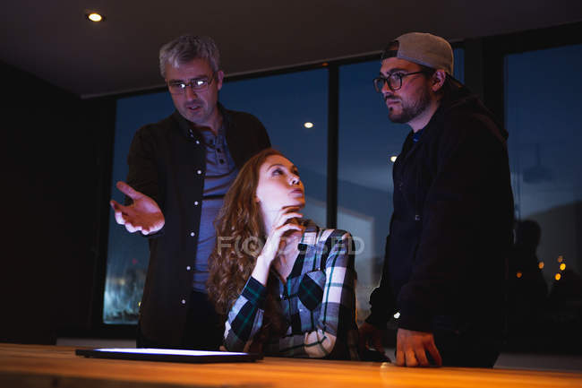 Vista frontal de una joven mujer caucásica y dos hombres caucásicos junto a una mesa en una creativa sala de conferencias de la oficina, utilizando una tableta en una discusión en una reunión - foto de stock