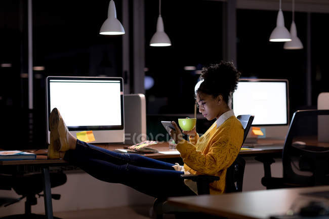 Vista lateral de una joven profesional de raza mixta que trabaja hasta tarde en una oficina moderna, sentada en un escritorio con los pies en alto usando una tableta y sosteniendo una taza - foto de stock