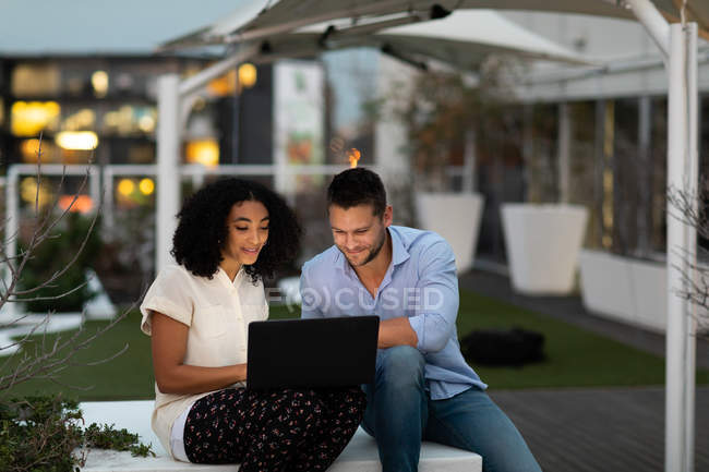 Vista frontal de un joven hombre profesional caucásico y una mujer de raza mixta que trabajan hasta tarde en una oficina moderna, sentados en la terraza de la azotea usando una computadora portátil juntos - foto de stock