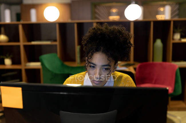 Vue de face d'une jeune femme de race mixte caucasienne travaillant tard dans un bureau moderne, assise à un bureau regardant un écran d'ordinateur de bureau — Photo de stock