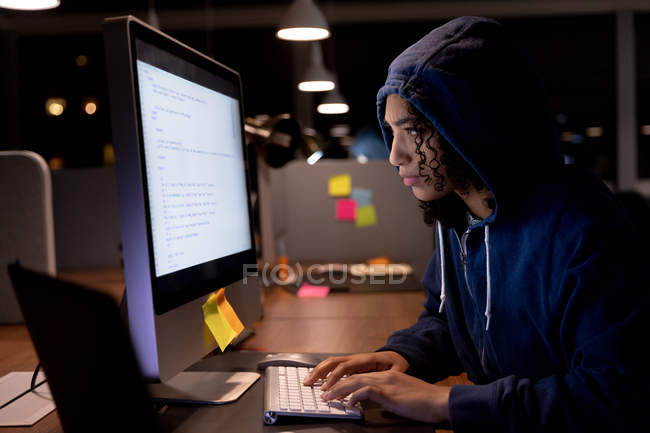 Side view of a young mixed race woman wearing a hoodie working late in a modern office, sitting at a desk using a desktop computer — Stock Photo