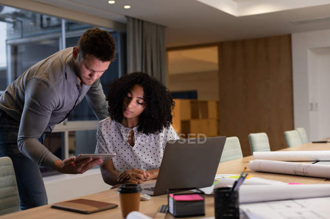 Frontansicht eines jungen kaukasischen Berufsmannes und einer Frau mit gemischter Rasse, die spät in einem modernen Büro am Schreibtisch arbeiten, einen Laptop benutzen und gemeinsam auf ein Tablet schauen — Stockfoto