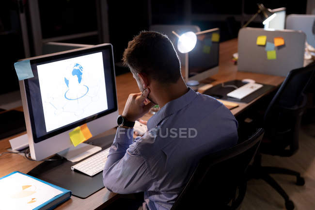 Vista trasera de un joven profesional caucásico que trabaja hasta tarde en una oficina moderna, sentado en un escritorio usando una computadora de escritorio - foto de stock