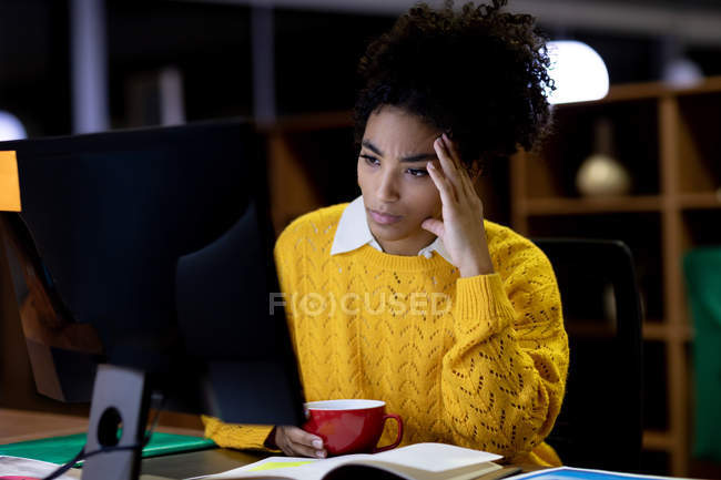 Front view of a young mixed race professional woman working late in a modern office, sitting at a desk, leaning and staring at a desktop computer monitor, holding a cup — Stock Photo