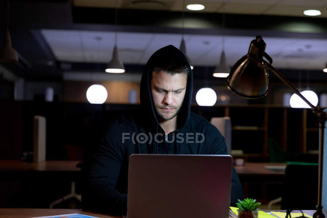 Frontansicht eines jungen kaukasischen Berufstätigen, der spät in einem modernen Büro arbeitet, am Schreibtisch sitzt und einen Kapuzenpullover mit einem Laptop benutzt — Stockfoto