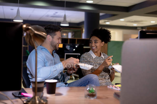 Front view of a young Caucasian professional man and mixed race woman working late in a modern office, sitting at a desk eating takeaway food and talking together — Stock Photo