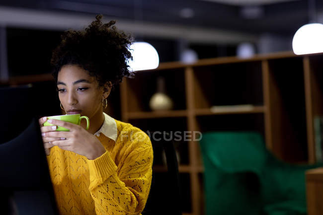 Frontansicht einer jungen Frau mit gemischter Rasse, die spät in einem modernen Büro arbeitet, an einem Schreibtisch sitzt, auf einen Desktop-Computermonitor blickt und eine Tasse in der Hand hält — Stockfoto