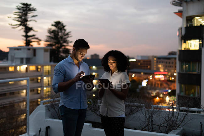 Front view of a young Caucasian professional man and mixed race woman working late at a modern office, standing on the roof terrace using tablet computers — Stock Photo