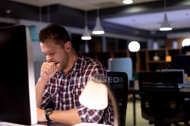 Vista frontal de un joven profesional caucásico que trabaja hasta tarde en una oficina moderna, sentado en un escritorio usando una computadora de escritorio y bostezando - foto de stock