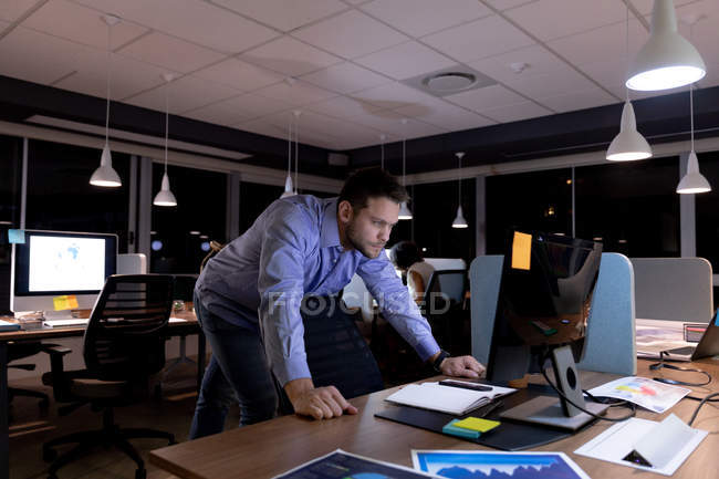 Вид спереди на молодого кавказского профессионала, работающего допоздна в современном офисе, стоящего за столом и смотрящего на монитор настольного компьютера — стоковое фото