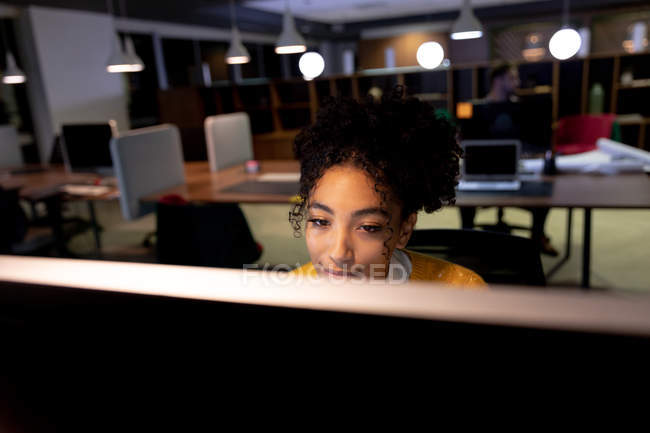 Вид спереди молодой профессиональной женщины смешанной расы, работающей допоздна в современном офисе, за монитором компьютера, сидящей за столом — стоковое фото