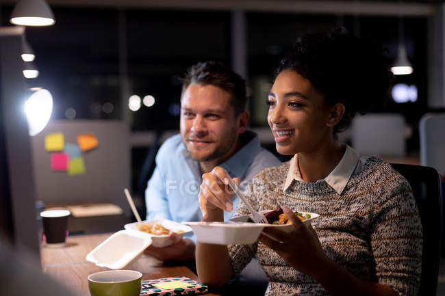 Frontansicht eines jungen kaukasischen Berufsmannes und einer Frau mit gemischter Rasse, die spät in einem modernen Büro arbeiten, an einem Schreibtisch sitzen, Essen zum Mitnehmen essen und gemeinsam lächelnd auf einen Computermonitor schauen — Stockfoto
