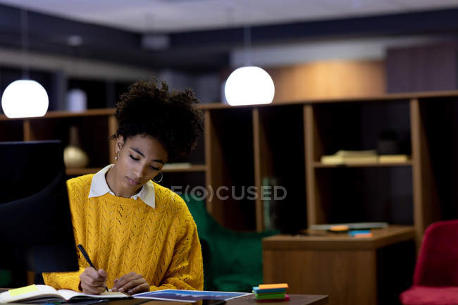 Vista frontal de una joven profesional de raza mixta que trabaja hasta tarde en una oficina moderna, sentada en un escritorio frente a un monitor de computadora tomando notas - foto de stock