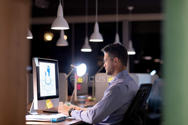 Vista lateral de un joven profesional caucásico que trabaja hasta tarde en una oficina moderna, sentado en un escritorio usando una computadora de escritorio y mirando fijamente al monitor - foto de stock