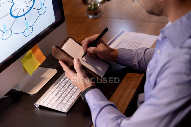 Vista lateral de perto de um jovem homem profissional caucasiano que trabalha até tarde em um escritório moderno, sentado em uma mesa usando um computador desktop e fazendo anotações — Fotografia de Stock
