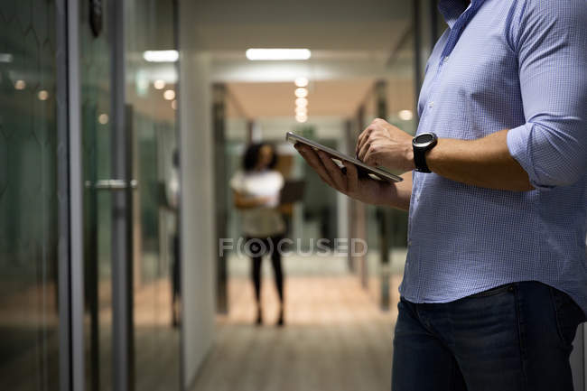 Side view mid section of a young Caucasian professional man and mixed race woman working late in a modern office, man standing in the corridor using a tablet computer and woman standing in the background — Stock Photo