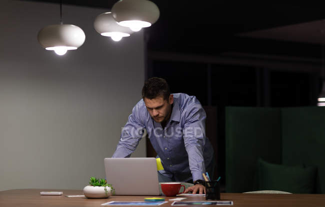 Frontansicht eines jungen kaukasischen Berufstätigen, der spät in einem modernen Büro arbeitet und mit einem Laptop am Schreibtisch steht — Stockfoto