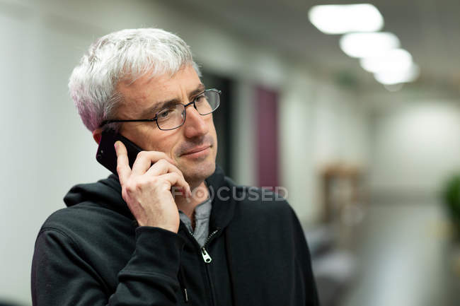 Vorderansicht eines kaukasischen Mannes mit grauen Haaren, der in einem kreativen Büro arbeitet, auf dem Smartphone spricht und eine Brille trägt — Stockfoto