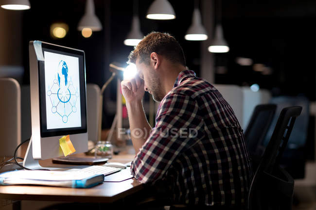 Vista lateral de um jovem homem profissional caucasiano trabalhando até tarde em um escritório moderno, sentado em uma mesa usando um computador desktop, inclinando a cabeça em sua mão — Fotografia de Stock