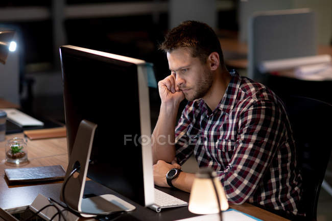 Бічний вид на молодого кавказького професіонала, який працював пізно в сучасному офісі, сидячи за столом за столом за допомогою настільного комп'ютера, схилившись і дивлячись на монітор. — стокове фото