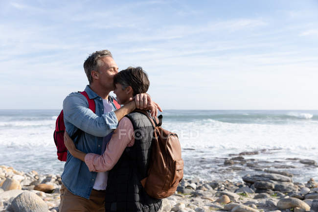 Vista frontal de um casal caucasiano adulto de meia idade desfrutando de tempo livre beijando ao lado do mar em um dia ensolarado — Fotografia de Stock