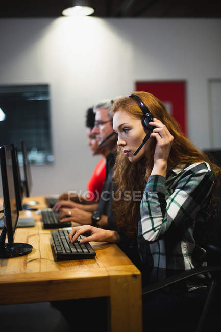 Vista lateral de uma jovem caucasiana em um escritório criativo, vestindo fone de ouvido com seus colegas no fundo. Isto é um negócio de arranque. — Fotografia de Stock