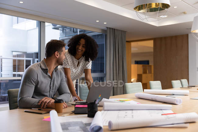 Vue de face d'un jeune homme professionnel caucasien et d'une femme métisse travaillant tard dans un bureau moderne à un bureau, utilisant un ordinateur portable et se souriant mutuellement — Photo de stock
