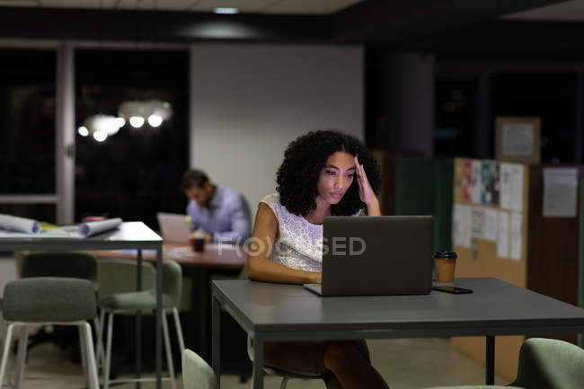 Vista frontal de uma jovem mulher profissional de raça mista e homem caucasiano trabalhando tarde em um escritório moderno, mulher sentada em uma mesa usando um computador portátil e homem sentado em segundo plano — Fotografia de Stock