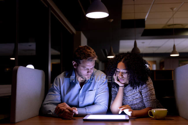 Vue de face d'un jeune homme professionnel caucasien et d'une femme de race mixte travaillant tard dans un bureau moderne assis à un bureau, regardant ensemble une tablette — Photo de stock
